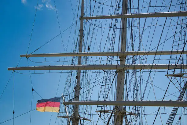 rigging and masts of a big sailing ship in front of a blue sky with the black-red-golden flag of the state Germany