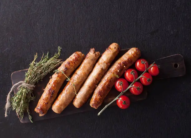 Photo of Fried sausages with thyme and tomatoes on a wooden serving Board. Great beer snack on a dark background. Top view with copy space