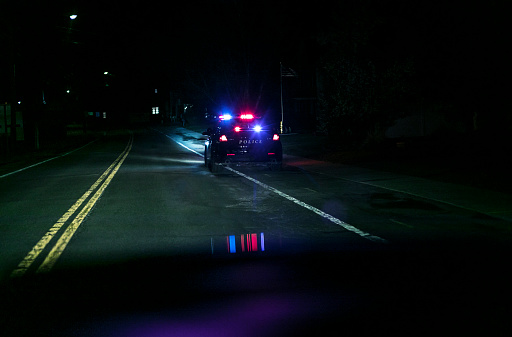 Car driver personal perspective POV at night from behind while approaching a stationary police car with flashing emergency lights at the edge of a rural road. A mostly obscured car waiting just ahead of the police cruiser has apparently been pulled over after a suspected traffic violation. Looks like someone has been caught in the act! (Vehicle details have been altered to make generic)