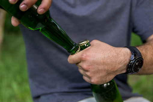 Opening beer bottles in nature without a beer opener, using one bottle to open another bottles, close up of male hands, nature picnic, holiday