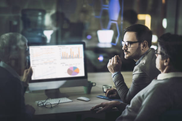 istock Business colleagues talking about charts while working on desktop PC at dark office. 1156312179