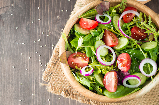 Fresh healthy vegetable salad made of cherry tomato, ruccola, arugula, feta, olives, cucumbers, onion and spices. Greek, Caesar salad in a white bowl on wooden background. Healthy organic diet food concept.