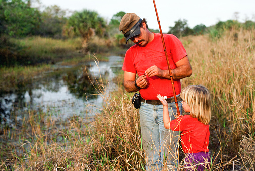 A horizontal image of a father and daughter fishing.  The father is wearing a hat and red t-shirt. The young toddler female is wearing a red t-shirt as well. The father is baiting the hook for his daughter.