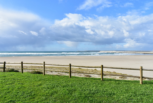 Sabon, A Coruña Province, Rias Altas, Galicia, Spain. Beach with grass and wooden fence. Industrial harbour and blue sea with waves and white foam. Blue sky with clouds.