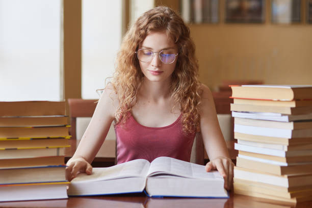 Portrait of hard working thoughtful college student with fair curly hair reading huge book, doing research, preparing for her lessons, looking attentively, having many tasks to do. Student life. retrato de estudiante universitario duro trabajando reflexivo con el pelo rizado justo leyendo enorme libro, haciendo investigación, preparándose para sus lecciones, mirando atentamente, teniendo muchas tareas que hacer. la vida estudiantil.