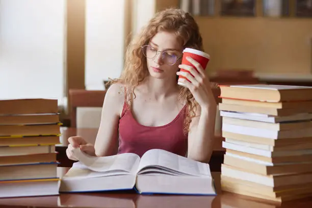 Tired determined college student searching for appropriate information for her project, reading texts, putting papercup of drink to her head, wearing red shirt and eyeglasses, working without stop.
