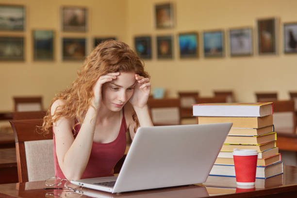 istock Task, test, learning and education concept. Intelligent, reader girl wearing casually sitting at desk in reading room in front of laptop, keeps hands on forehead, preparing for important exam. 1156331092