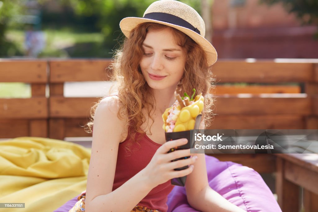 Young woman sitting on farameless chair in park, holding in hands ice cream, wearing burgundy casual t shirt and hat, looks mysteriously down, enjoying to spend summer day outdoor. Recreation concept. Activity Stock Photo