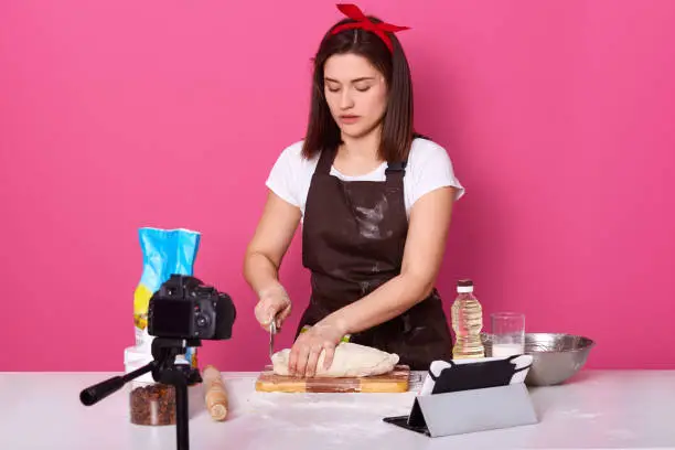 Photo of Hard working creative cook wearing red headband, brown apron and white t shirt cutting raw pie, following recipe, recording cooking tutorial for subscribers, busy with pie, looking concentrated.