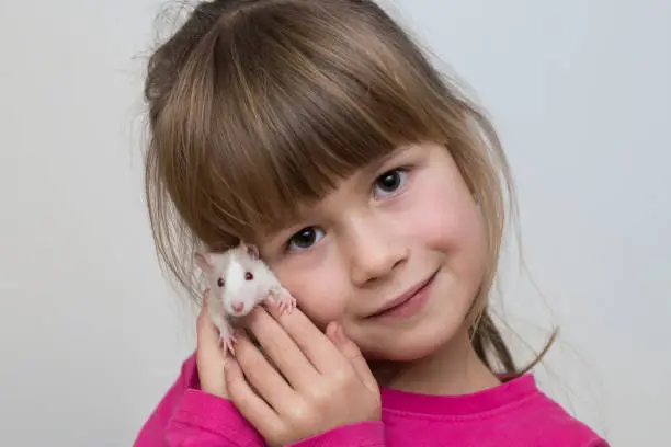Photo of Portrait of happy smiling cute child girl with white pet mouse hamster on light copy space background. Keeping pets at home, care and love to animals concept.