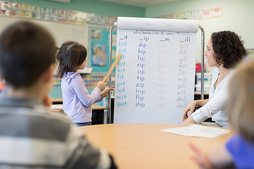 A first grader stands in front of a giant hanging sheet of spelling words and sounds them out in front of the class while she points to them with a ruler. The teacher, an female, is encouraging her.