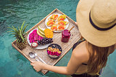 Breakfast tray in swimming pool, floating breakfast in luxury hotel. Girl relaxing in the pool drinking smoothies and eating fruit plate, smoothie bowl by the hotel pool. Exotic summer diet. Tropical beach lifestyle. Bali Trend