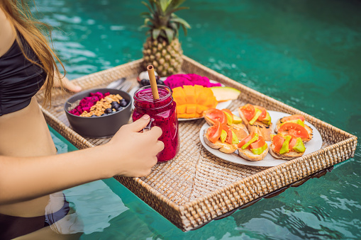 Breakfast tray in swimming pool, floating breakfast in luxury hotel. Girl relaxing in the pool drinking smoothies and eating fruit plate, smoothie bowl by the hotel pool. Exotic summer diet. Tropical beach lifestyle. Bali Trend.