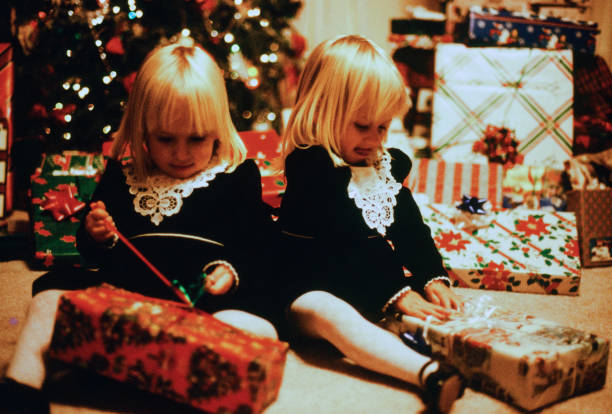 A horizontal image taken indoors of two blonde girls opening up Christmas packages. They are wearing matching black dresses with white lace. The Christmas tree and additional packages are in the background. The girls are engaged in unwrapping their gifts.