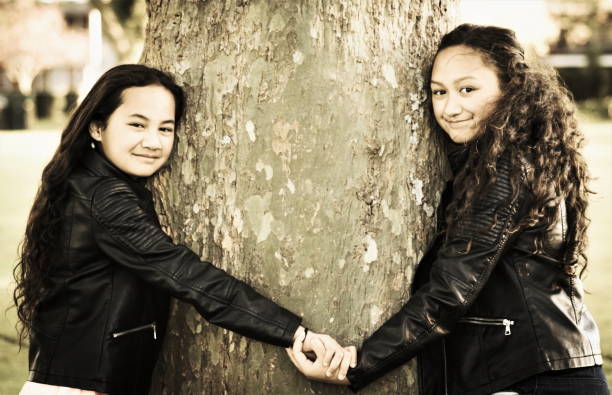 Two Maori sisters holding hands hugging a tree outdoors in the day