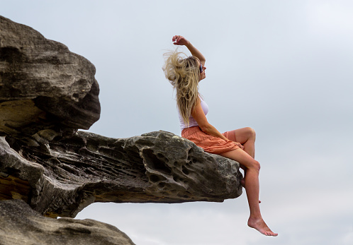 Happy carefree woman tossing her hair whilst sitting on a narrow rock jutting out from the main cliff line.