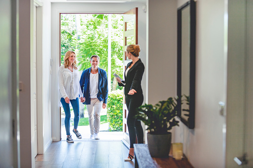 Real estate agent showing a young couple a new house. The house is contemporary. All are happy and smiling. The couple are casually dressed and the agent is in a suit. The couple are being greeted at the front door by the real estate agent. Copy space