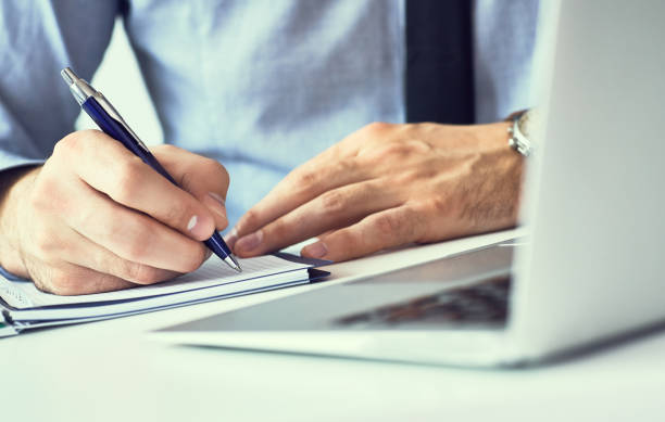istock Businessman hand writing note on a notebook. Business man working at office desk. Close up of empty notebook on a blackboard with office supplies. 1156396231