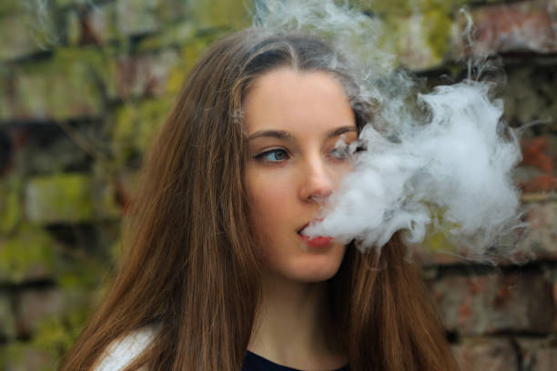 Vape teenager. Young pretty white girl in white cardigan smoking an electronic cigarette opposite destroyed brick wall on the street in the spring. Bad habit. Vape teenager. Young pretty white girl in white cardigan smoking an electronic cigarette opposite destroyed brick wall on the street in the spring. Bad habit that is harmful to health. electronic cigarette stock pictures, royalty-free photos & images