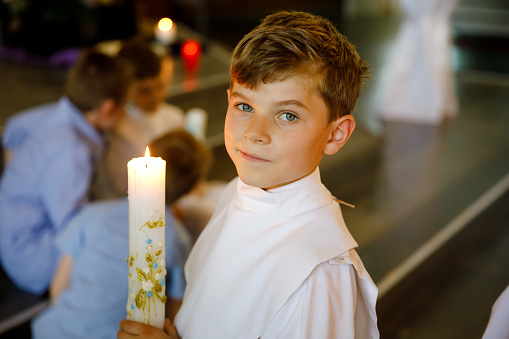 Little kid boy receiving his first holy communion. Happy child holding Christening candle. Tradition in catholic curch. Kid in a white traditional gown in a church near altar