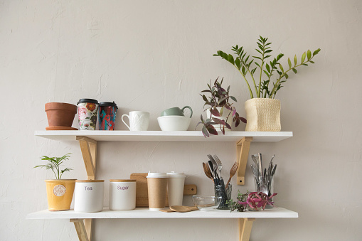 Nice shelves with various tableware and ceramic pots hanging on white wall in stylish room