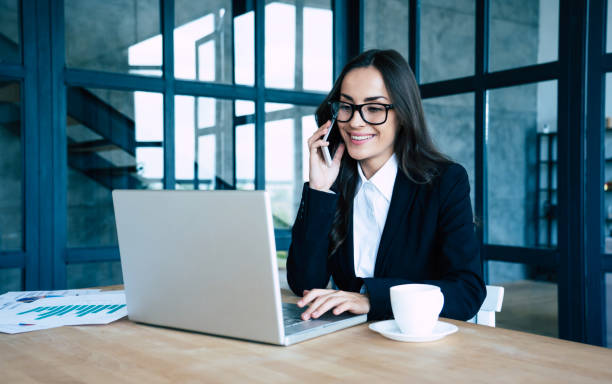 istock Charming happy business women dressed in full suit using smartphone and laptop while sitting in modern office 1156432232
