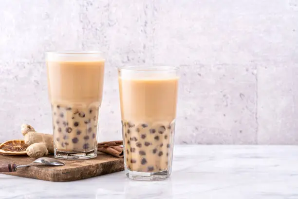 Photo of Popular Taiwan drink - Bubble milk tea with tapioca pearl ball in drinking glass on marble white table wooden tray background, close up, copy space