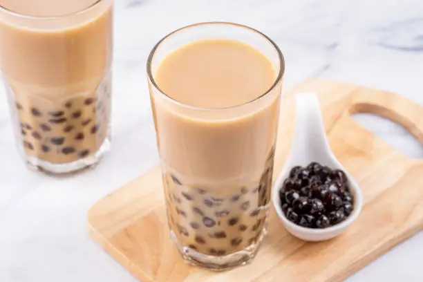 Photo of Popular Taiwan drink - Bubble milk tea with tapioca pearl ball in drinking glass on marble white table wooden tray background, close up, copy space