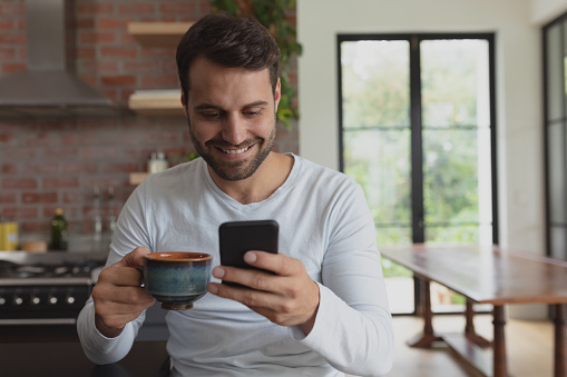 Front view of happy Caucasian man with coffee cup using mobile phone in a comfortable home