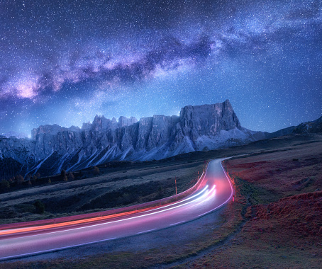 Milky Way over mountain road at night in summer. Blurred car headlights on winding road. Colorful landscape with sky with stars and purple milky way, light trails, rocks, trees and highway. Space