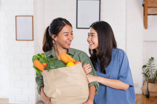 lésbica asiática feliz dos pares que prende o saco vegetal após a compra na mercearia em casa. conceito de estilo de vida lgbtq. - bff - fotografias e filmes do acervo