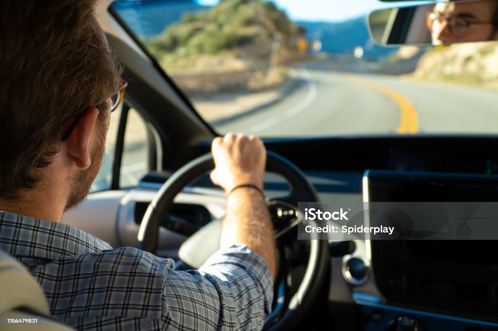 Young Man Driving His New Hydrogen Car Close up of millennial young man at the steering wheel loving life and his new alternative energy car. He is on a road trip through the mountains. We see his reflection in the rear view mirror. Electric Vehicle Stock Photo