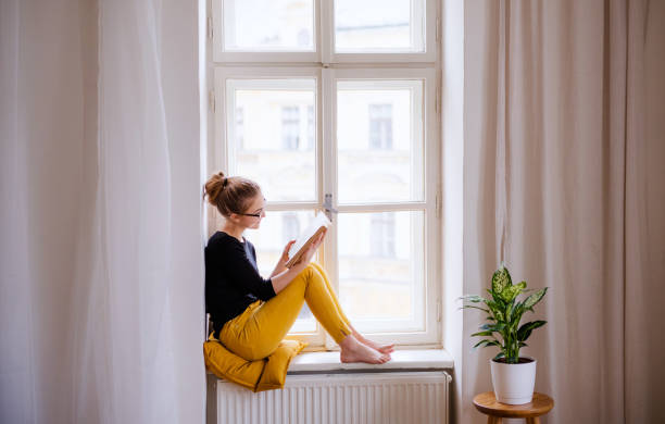 A young female student with a book sitting on window sill, studying. A young happy college female student with a book sitting on window sill at home, studying. reading stock pictures, royalty-free photos & images