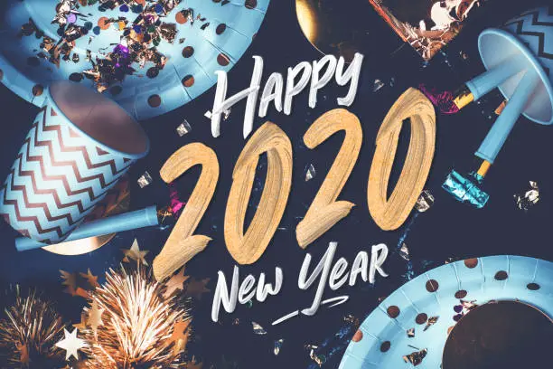 Photo of 2020 happy new year hand brush storke font on marble table with party cup,party blower,tinsel,confetti.Fun Celebrate holiday party time table top view.blue modern tone filter.festive greeting card.