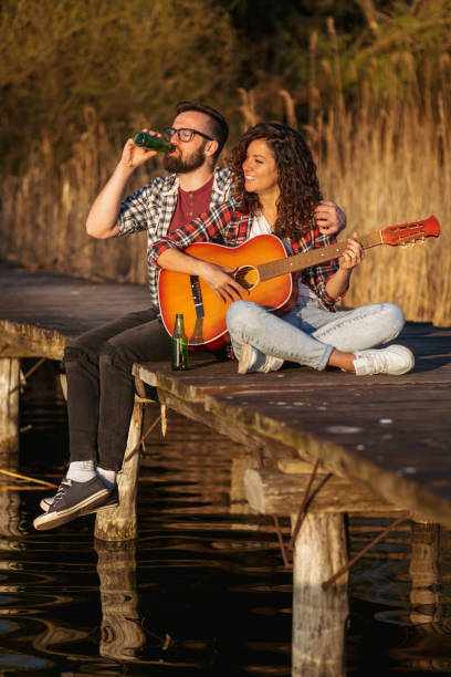 Couple sitting at the lake docks, playing the guitar, singing and drinking beer, enjoying a day in nature and relaxing