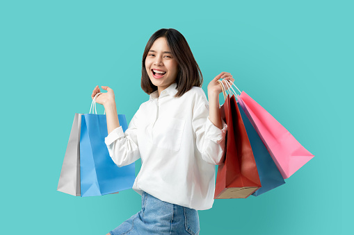 Portrait of young smiling asian woman casual clothes holding multicolored shopping bags on light blue background.