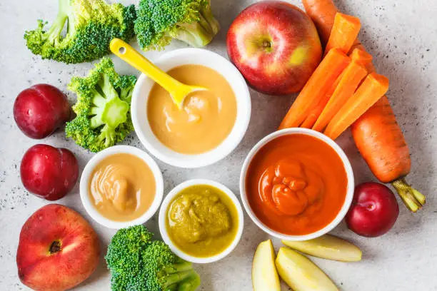 Vegetable and fruit baby puree (apple, broccoli, carrot, plum) in white bowl with ingredients. Baby food concept.