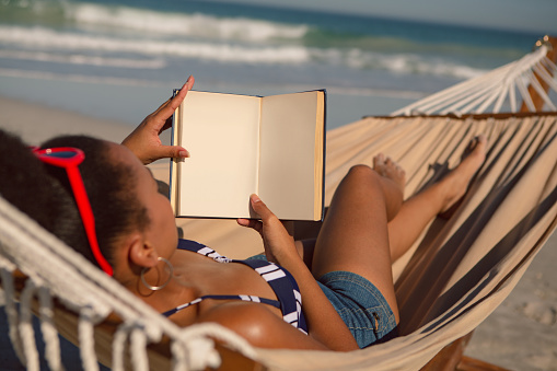 Rear view of African american woman reading a book while relaxing on hammock on beach in the sunshine