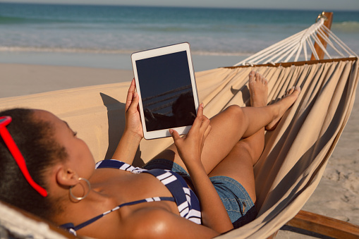 Rear view of African american woman using digital tablet while relaxing on hammock on beach in the sunshine
