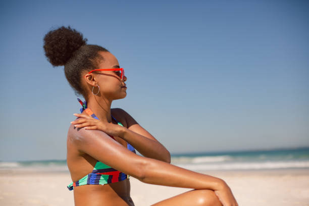 woman in bikini applying sunscreen lotion on shoulder at beach in the sunshine - hair bun hairstyle beautiful looking imagens e fotografias de stock