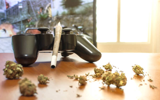 istock Video games and cannabis. Video game controller, big joint and marijuana buds on wooden table. Blur background of tv with video game and bright window 1156509878