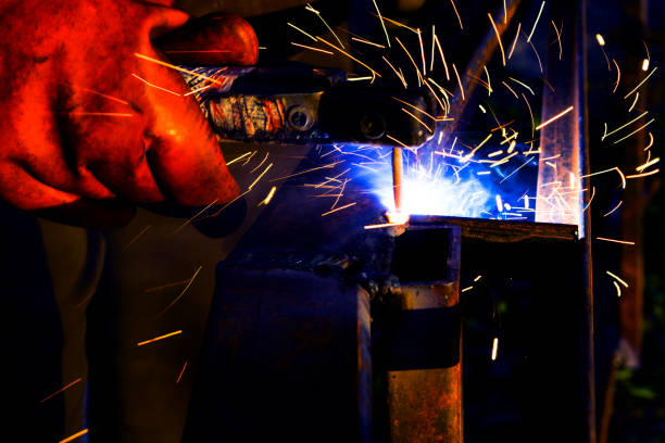 istock Welder worker performs jump welding. Man welder in protective gloves performs arc-welding process of metal structures. Flying sparks from the welding machine. Background construction site 1156512254