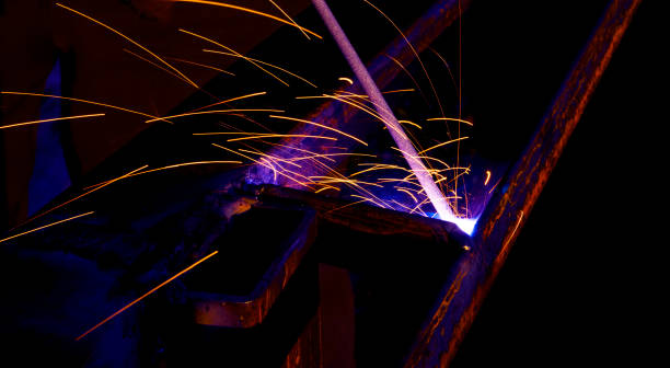 istock Welder worker performs jump welding. Man welder in protective gloves performs arc-welding process of metal structures. Flying sparks from the welding machine. Background construction site 1156512478