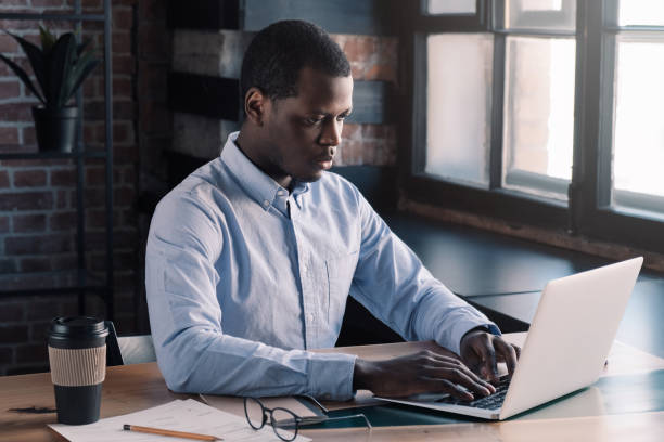 istock Young african american business man working, using laptop at office while sitting at table with papers and cup of coffee, typing message on computer keyboard 1156514362
