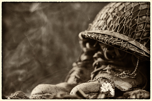 A vintage looking photo of a close-up of a pair of muddy military dog tags sitting on a pair of muddy boots with an army helmet sitting on top. This is a makeshift memorial for a fallen soldier on the battlefield. This could be from World War II or the Korean war.