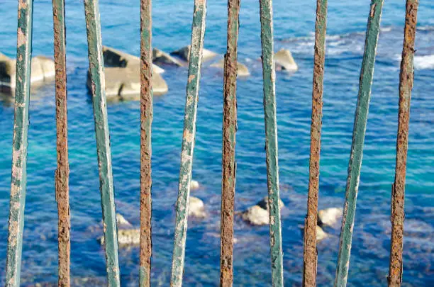 A row of rusted turquoise iron railings with peeling paint; the sunlit rocky sea visible between them.