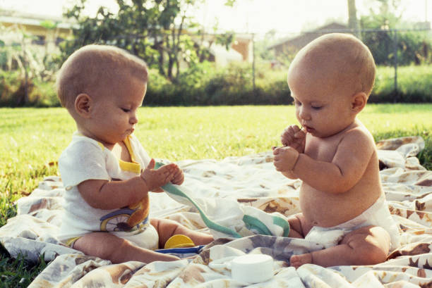 A horizontal image shows 2 female infants about 6 months of age.  They are sitting, facing each other, but are engrossed in their hands. It is a sunny day and they are sitting in the shade.