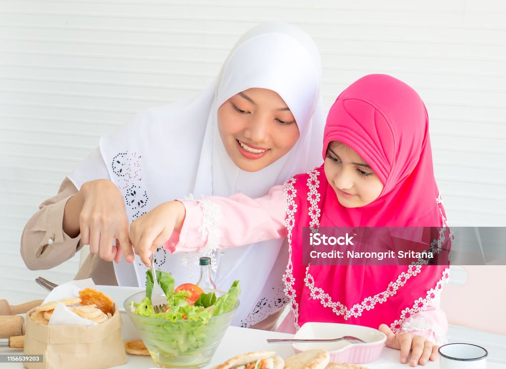 Muslim mother point to vegetable salad while little girl with pink hijab has fun with mixing salad put on the table Adult Stock Photo