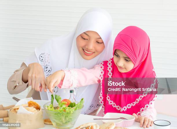 Muslim Mother Point To Vegetable Salad While Little Girl With Pink Hijab Has Fun With Mixing Salad Put On The Table Stock Photo - Download Image Now