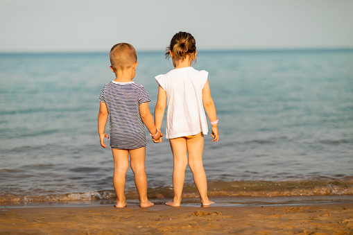 Cute little baby boy and a girl standing on a beach and holding hands. Children enjoying beach life on a sunny summer day